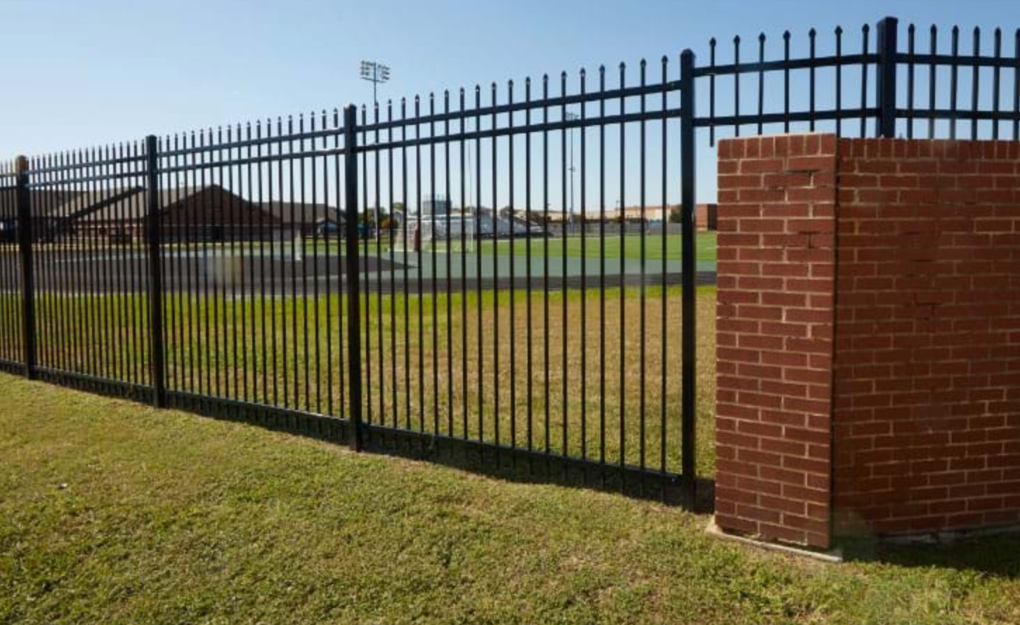 Black metal fence bordering a grassy area and a brick wall. In the background is a green field and a building.