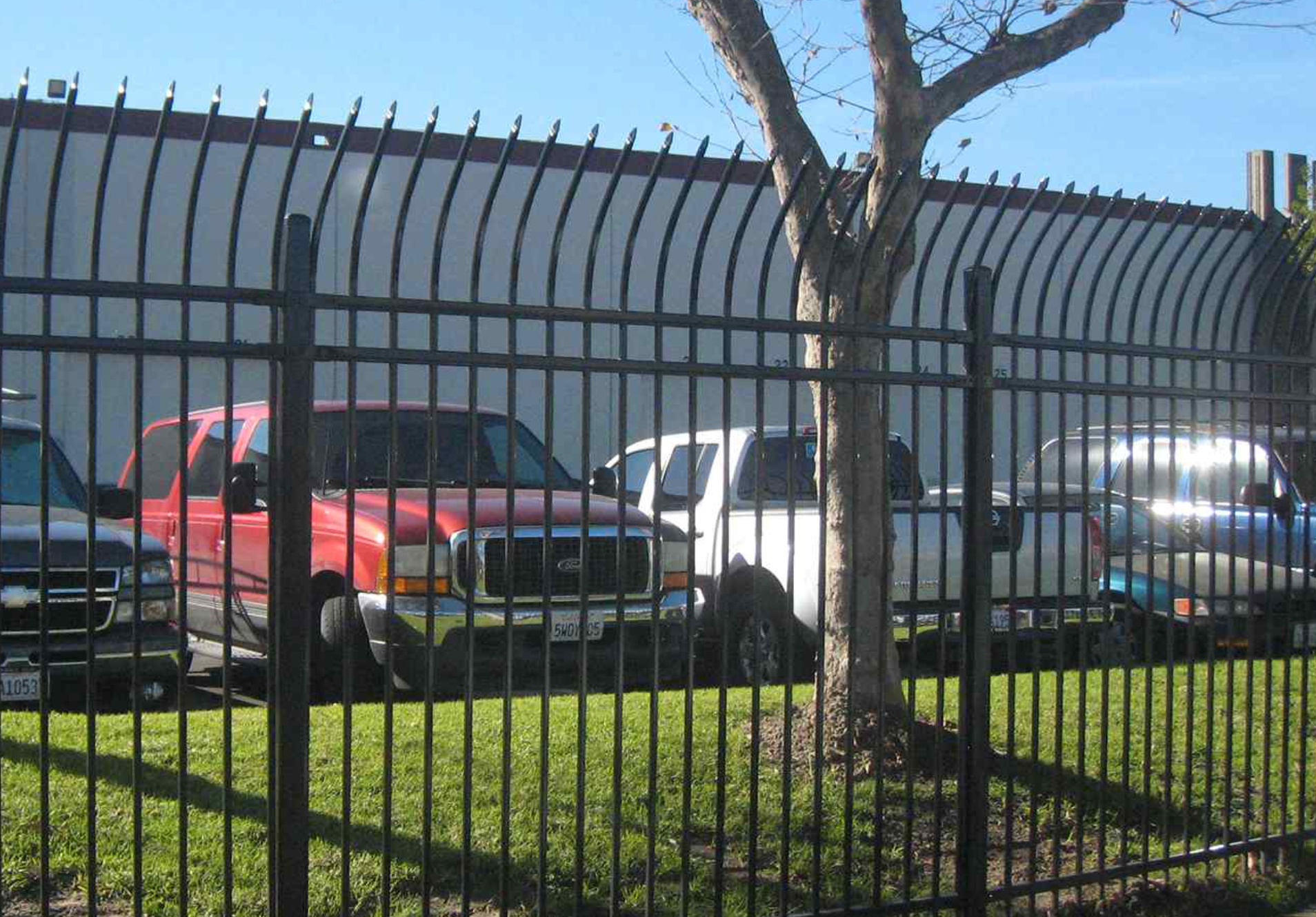 Black fence in front of several parked trucks in front of a tan building under a blue sky.