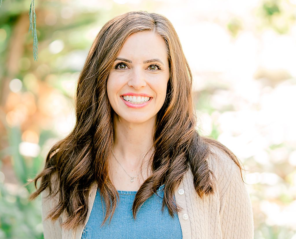 Woman with long brown hair smiles at the camera, wearing a blue top and tan cardigan outdoors.