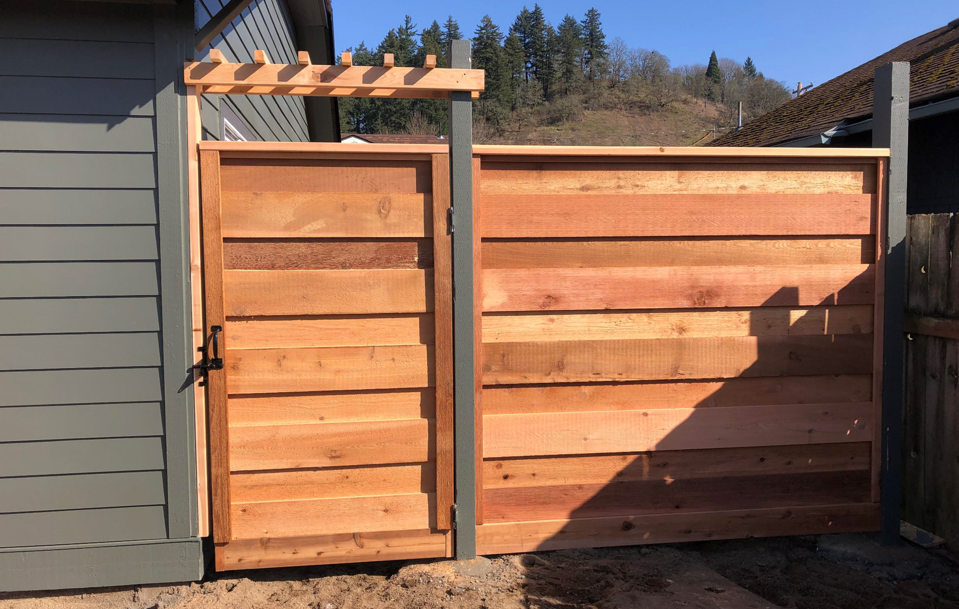 Wooden fence with a gate, in front of a building and trees under a sunny sky.