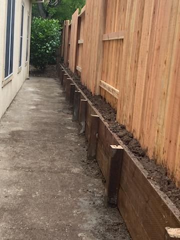 Narrow concrete walkway beside a house and wooden fence, with raised soil beds along the fence