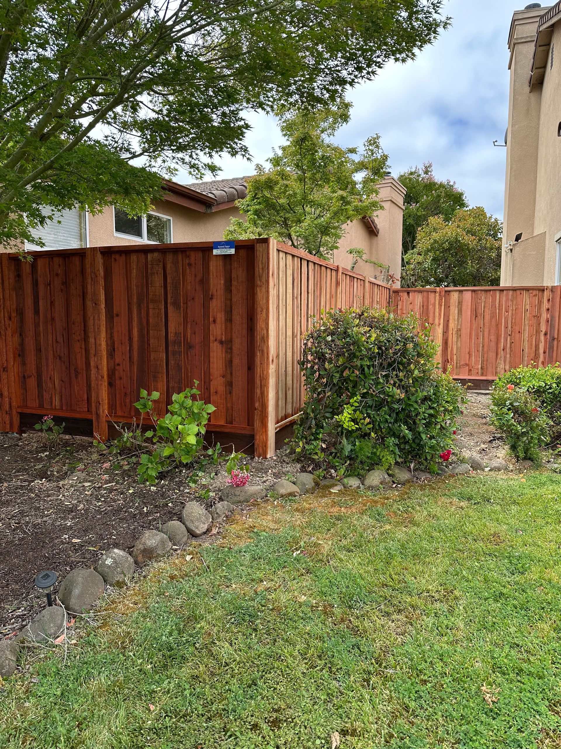 Wooden fence and shrubs along a grassy yard beside a stucco house under trees