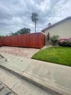 Residential street with red wooden fence, gravel strip, and green lawn beside a concrete sidewalk.