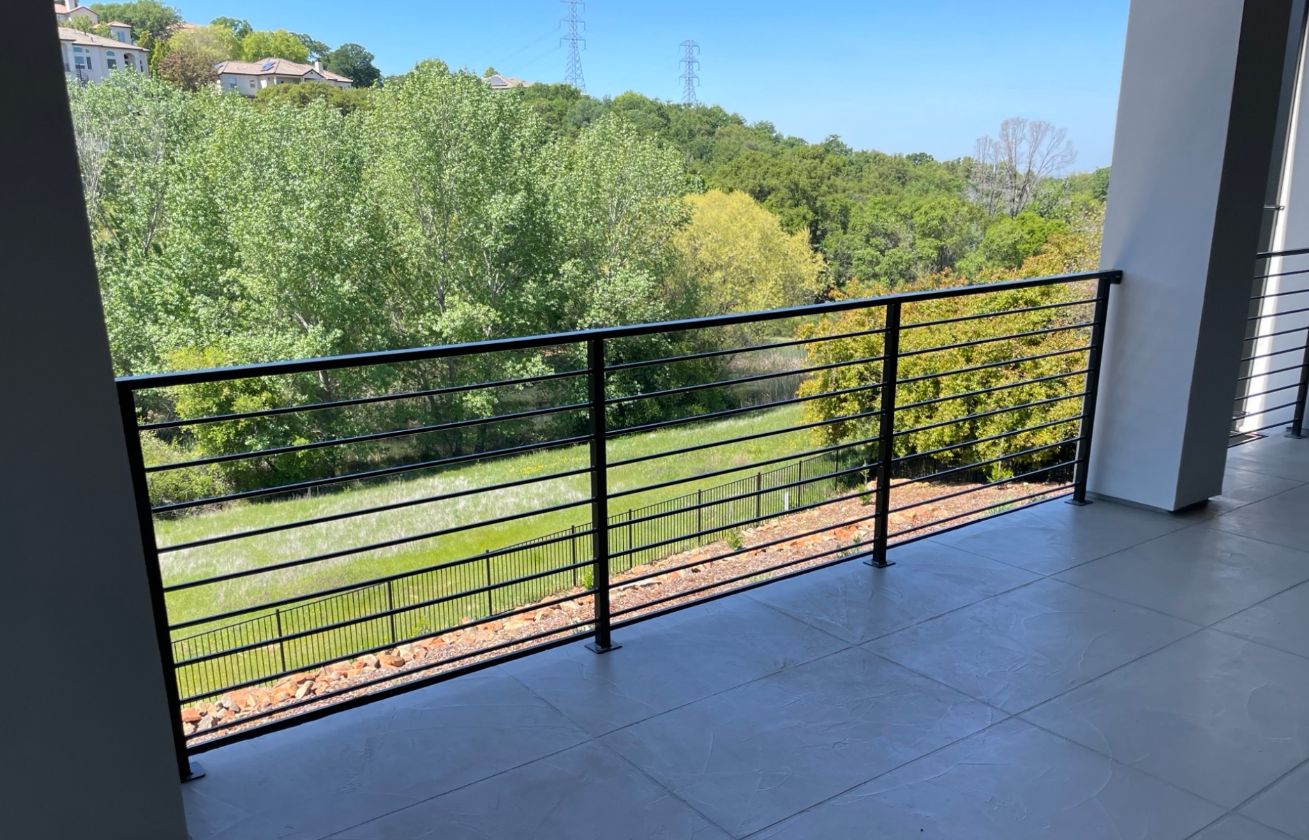 Balcony with black railing, overlooking green trees and grass on a sunny day.