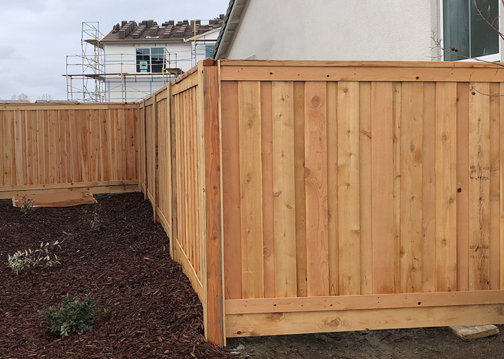 Wooden fence surrounding a yard with brown mulch and a house under construction in the background.
