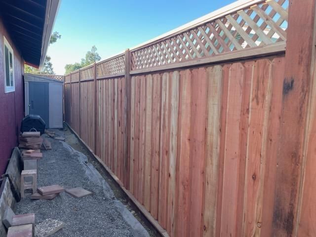 Narrow side yard with a tall wooden fence and lattice top beside a house, with gravel path and gate at the end.