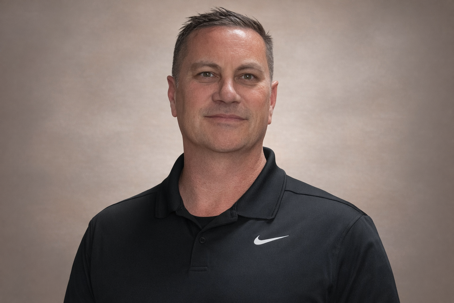 Man in black Nike polo shirt smiles. Neutral background.