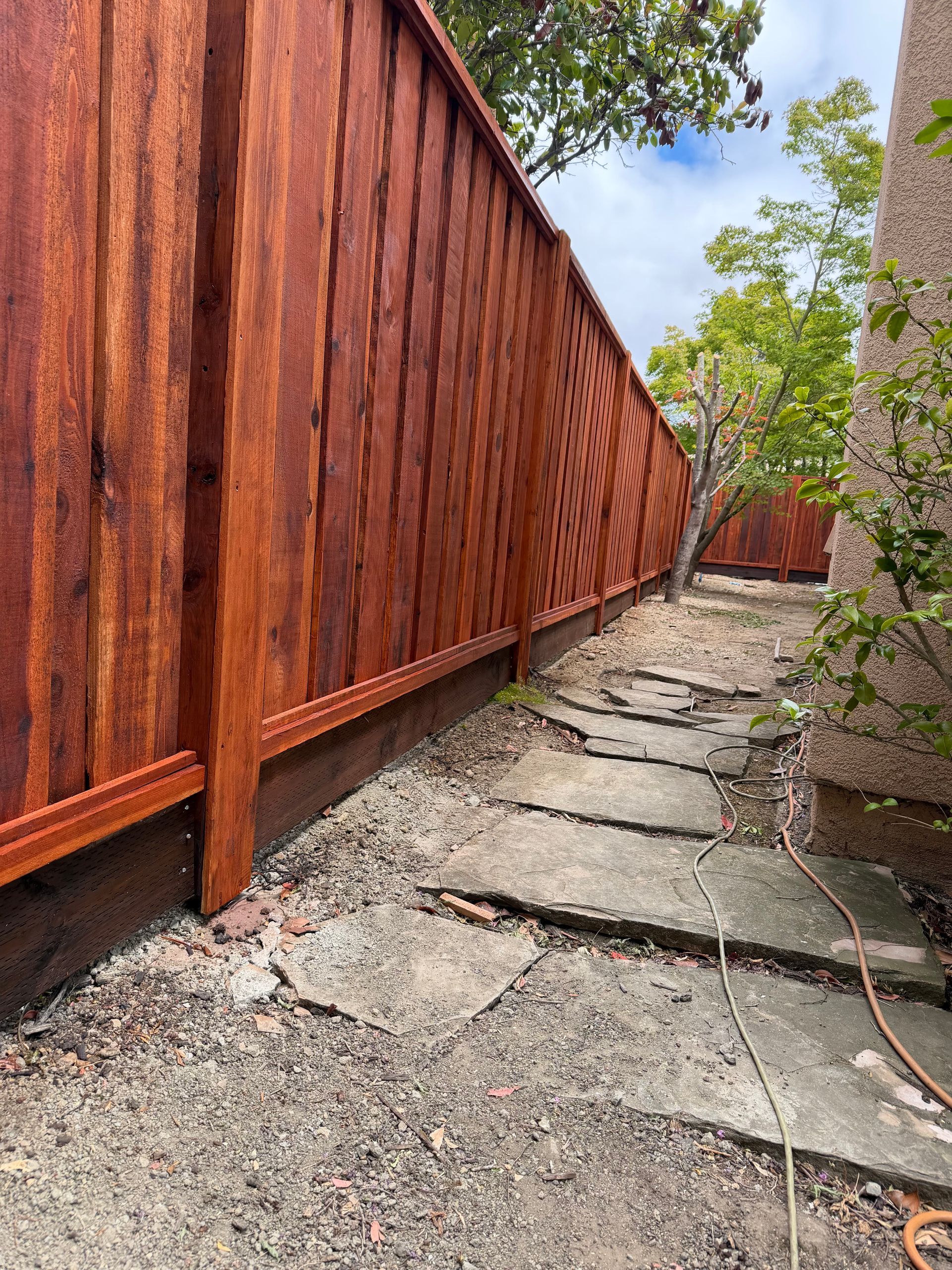 Narrow concrete walkway beside a tall red wooden fence, with trees and a small garden on the right.