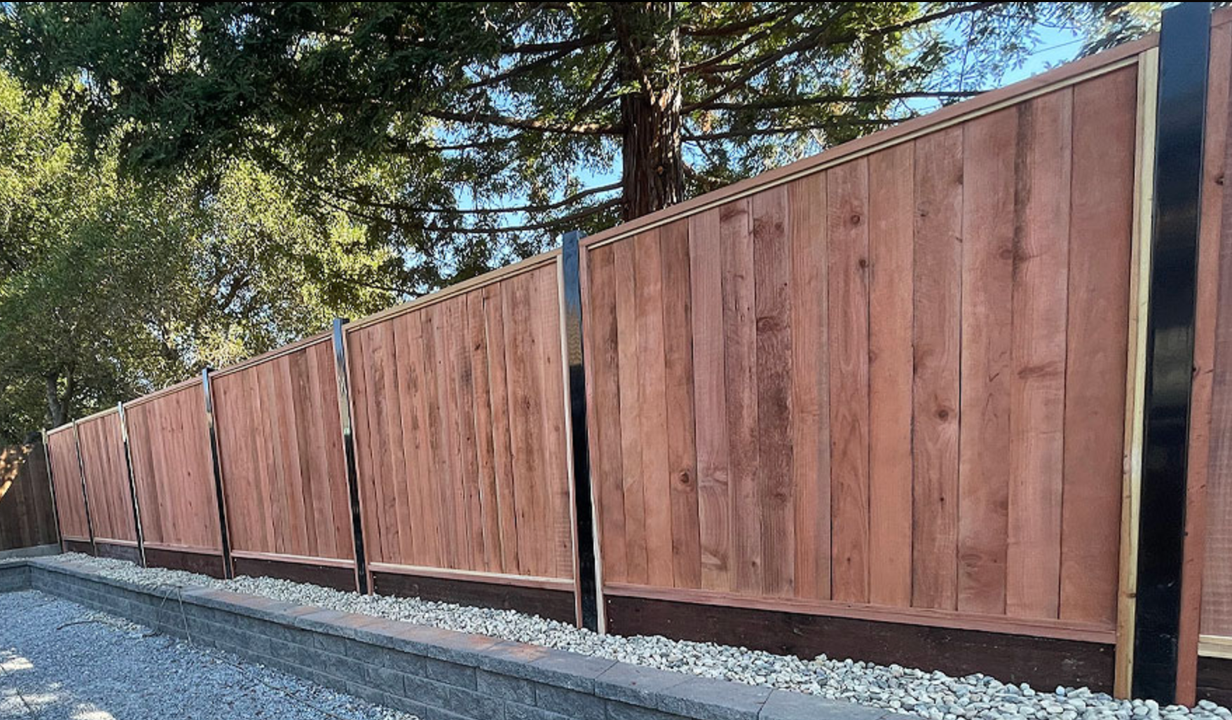 Wooden fence with metal posts along a gravel driveway, trees in the background.