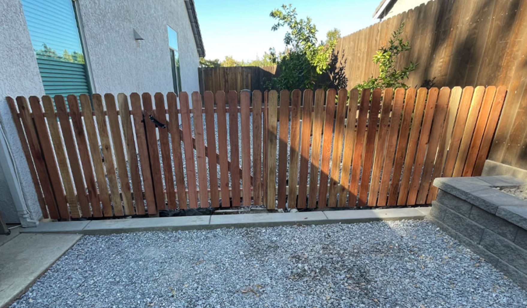 Wooden fence with gate in gravel area, next to a building and a retaining wall.