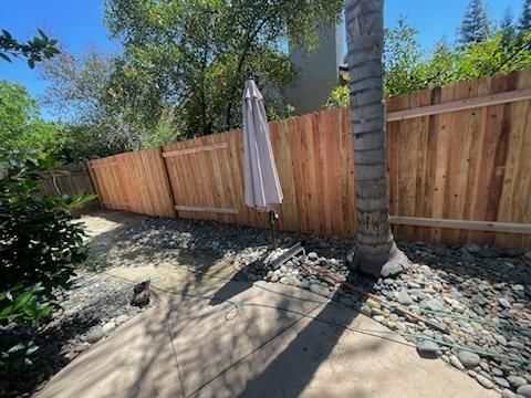 Sunny backyard patio with wooden fence, umbrella, palm tree, and stone landscaping.