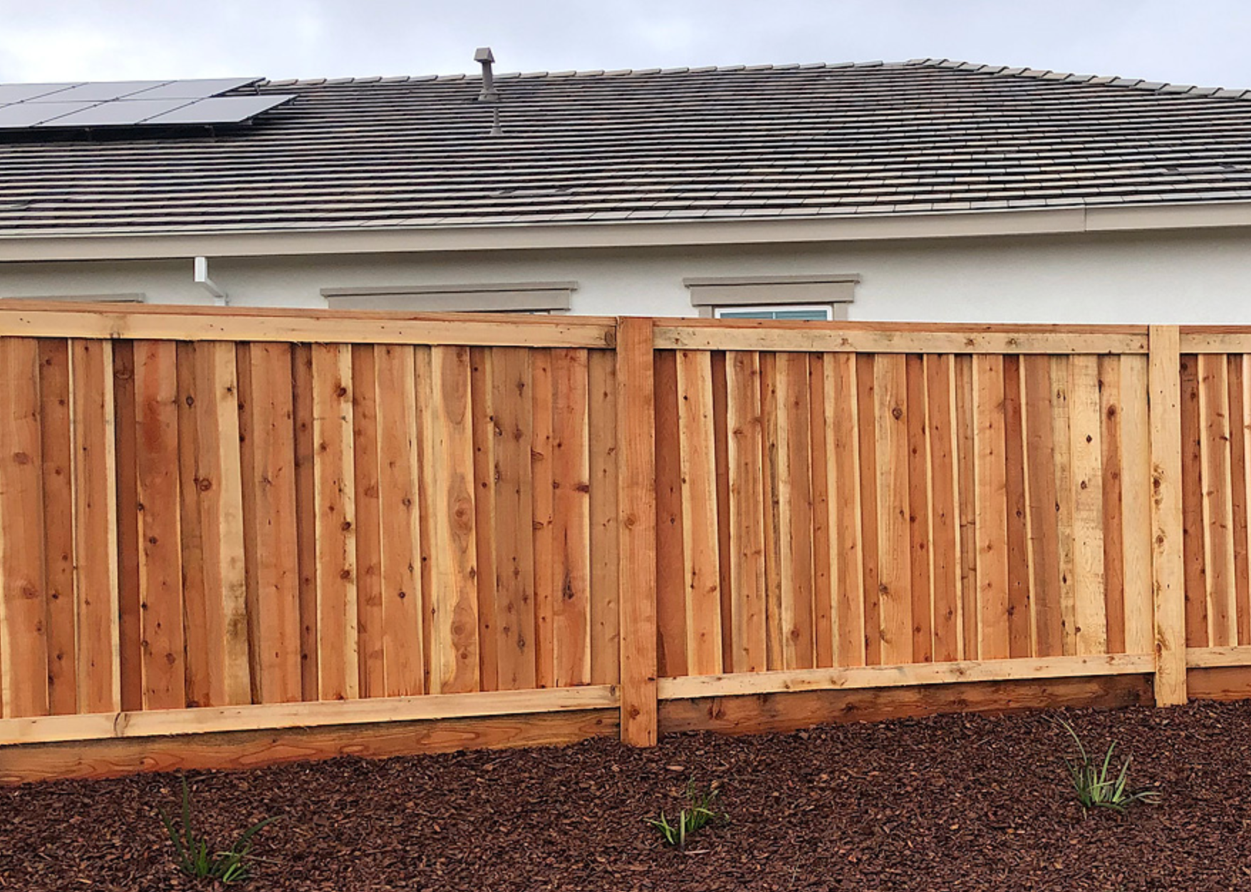 Wooden fence in front of a house, set in brown gravel. The roof is visible.