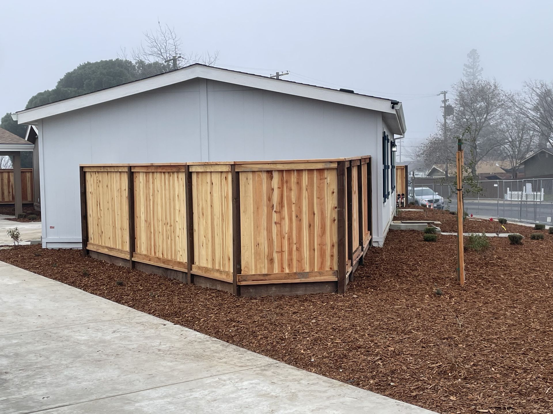 A light blue building with a wooden fence. Ground covered in brown mulch. Overcast day.