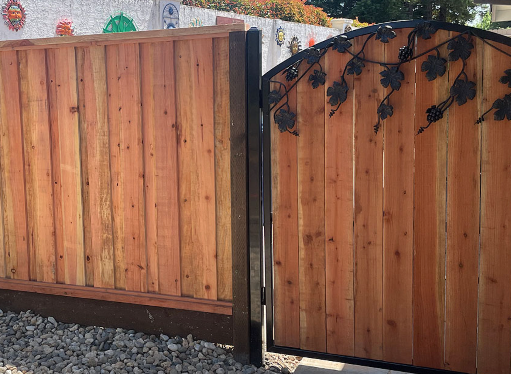 Wooden fence with gate, adorned with wrought iron leaves, set in a gravel area.