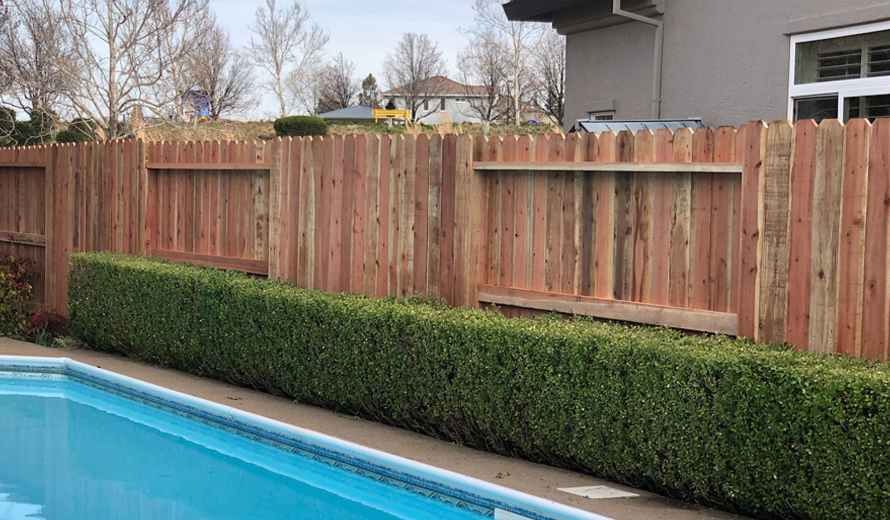Poolside view with wooden fence, hedge, and clear blue water.