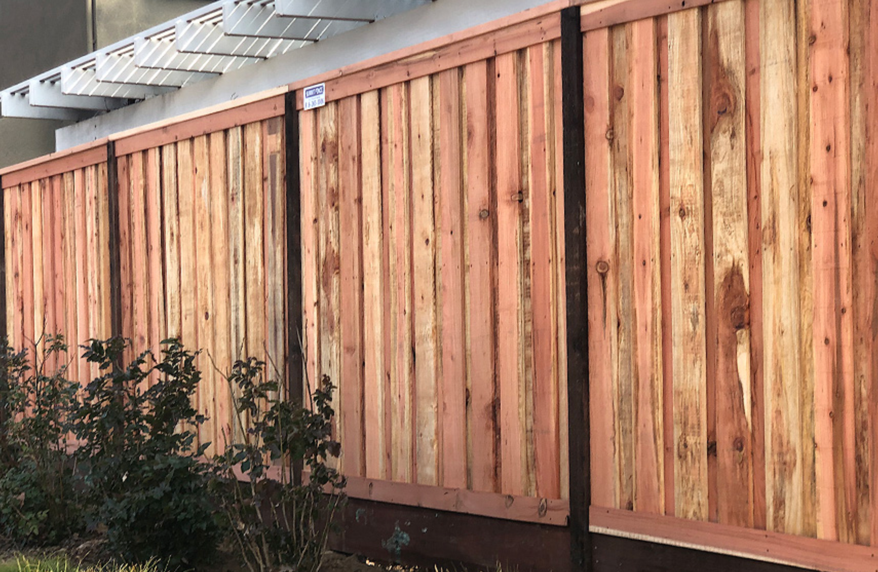 Wooden fence with vertical planks, brown and tan, near a building and shrubs.