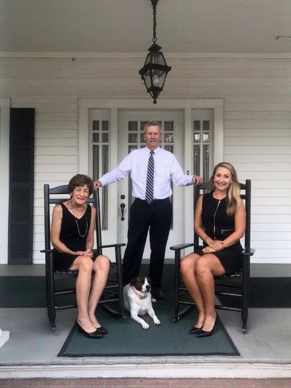 Three people and a dog pose on a porch. A man in a suit stands between two women in black dresses, with a dog at their feet.