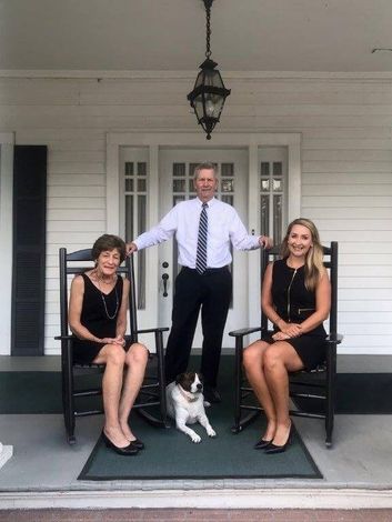 Three people and a dog pose on a porch. A man in a suit stands between two women in black dresses, with a dog at their feet.