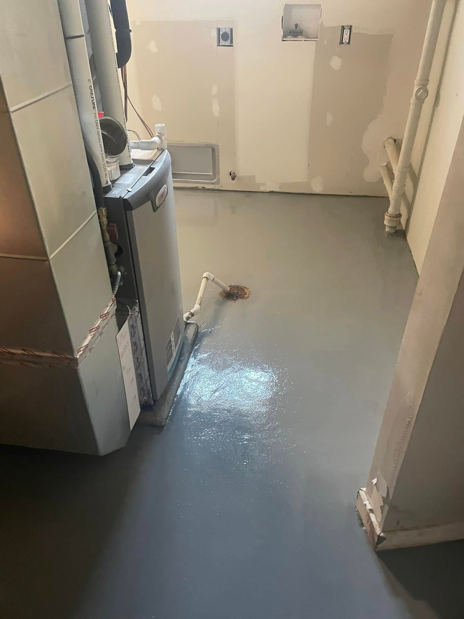 A gray, epoxy-coated floor in a utility room with a furnace unit and a floor drain with a small amount of pooled water.