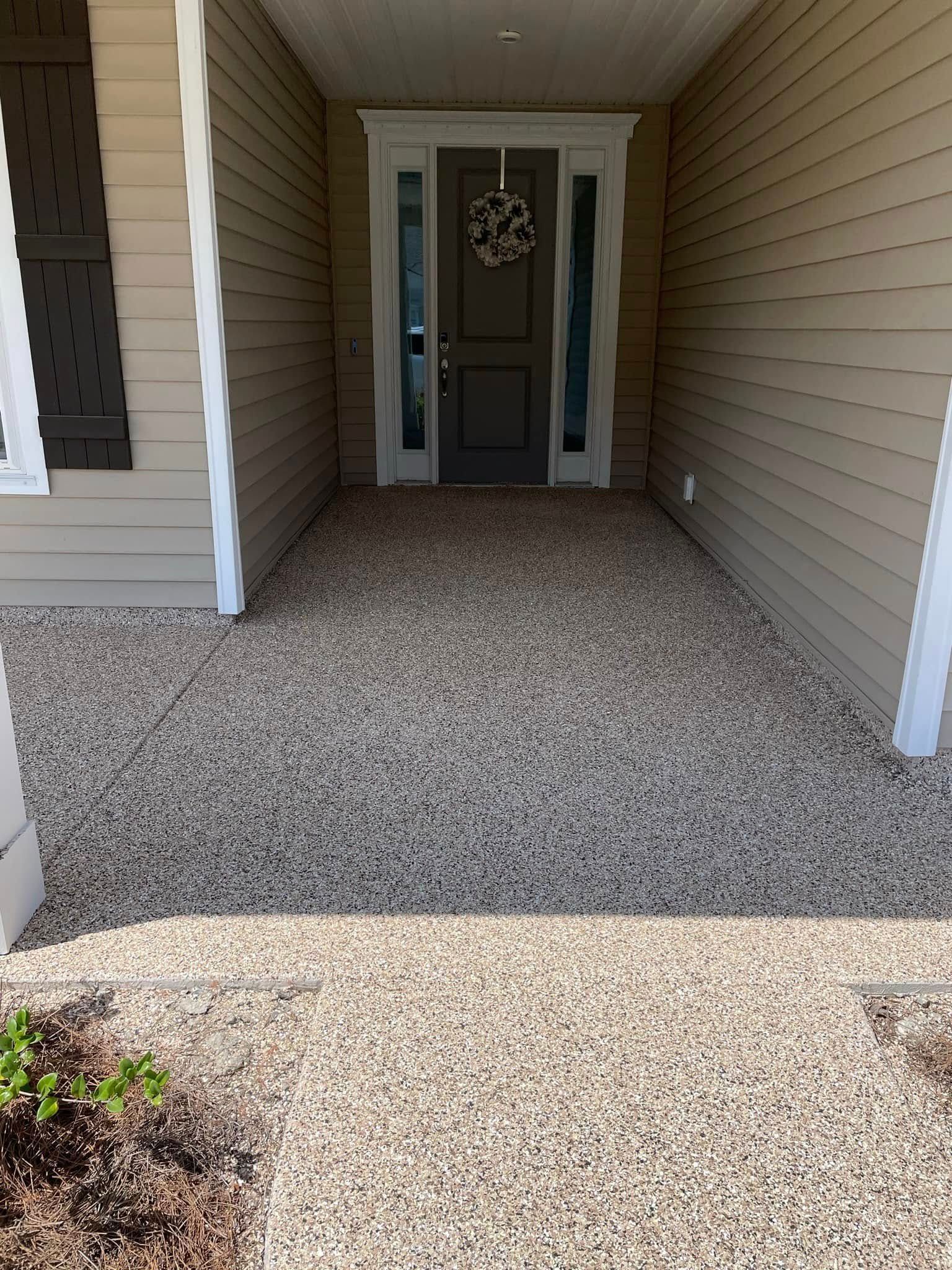 A tan house entrance with a gray front door, side windows, a wreath, and a light gravel porch floor.