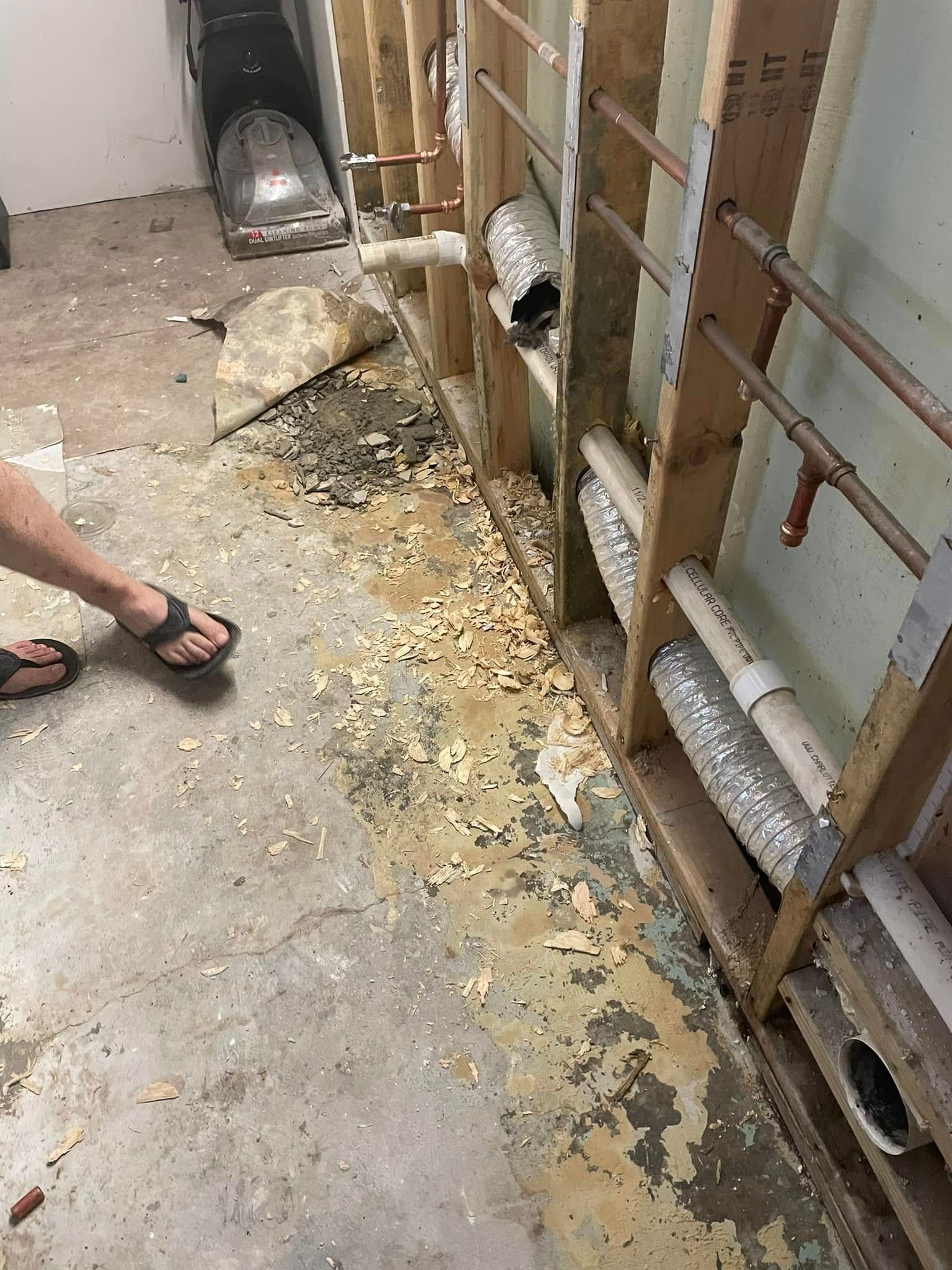 A construction site showing open wall studs with exposed plumbing and wiring over a debris-filled concrete floor.