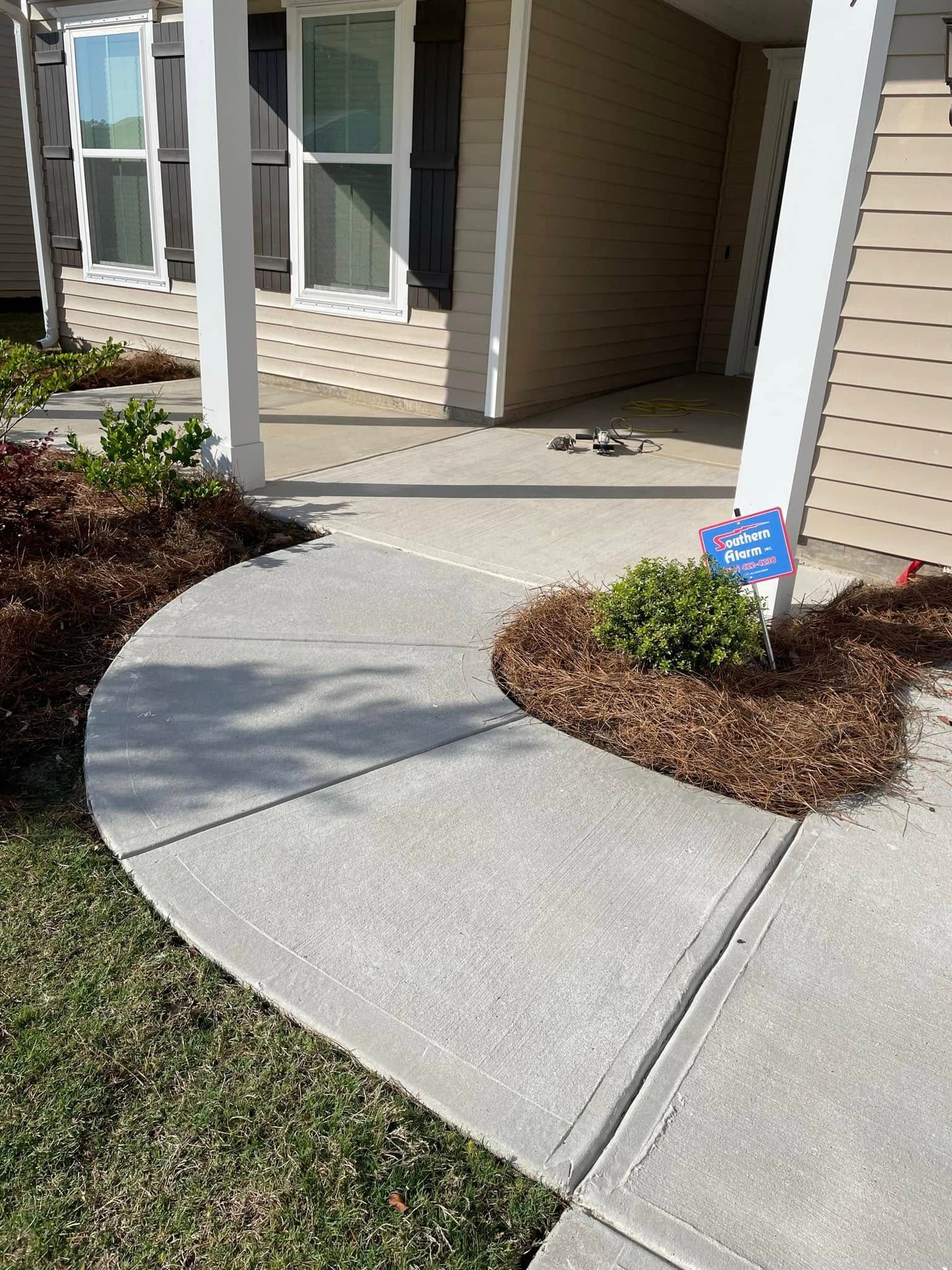 A concrete walkway curves toward the covered porch of a tan house with dark shutters and a small bush in mulch.