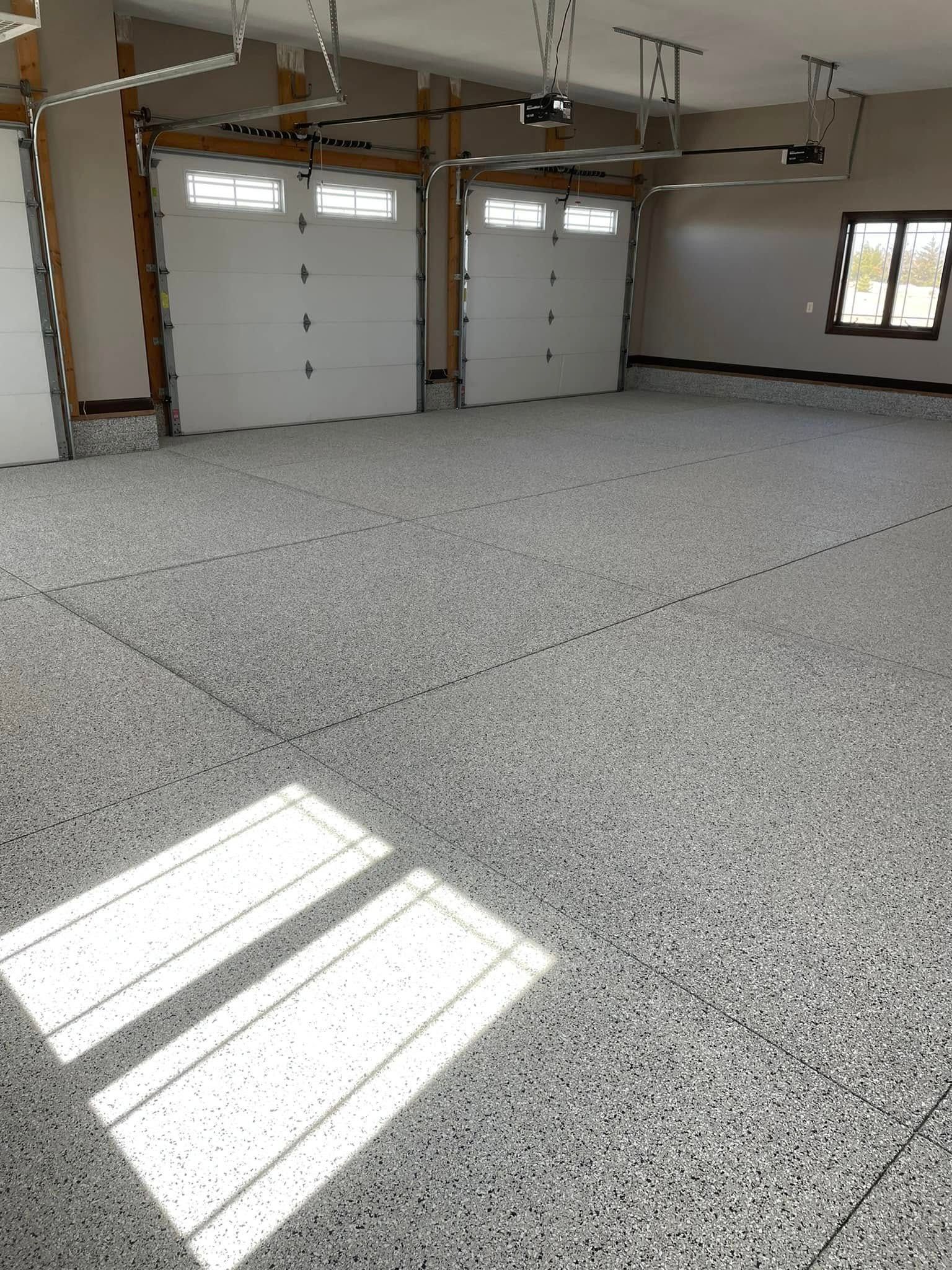 A spacious garage featuring a gray speckled epoxy floor, two white garage doors, and a window on the far wall.