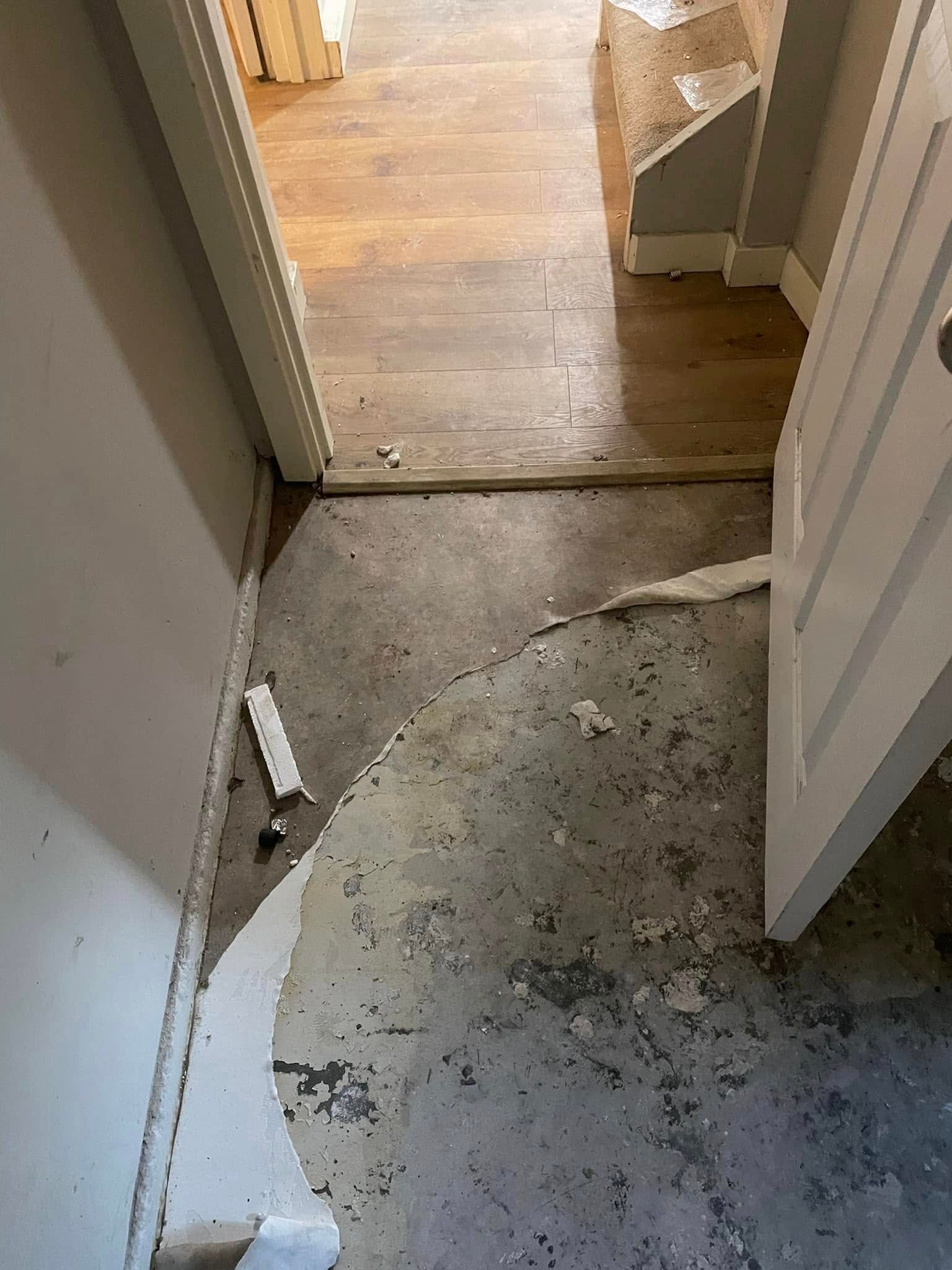 A doorway showing a transition from a room with damaged, exposed flooring to an adjacent room with finished wood floors.
