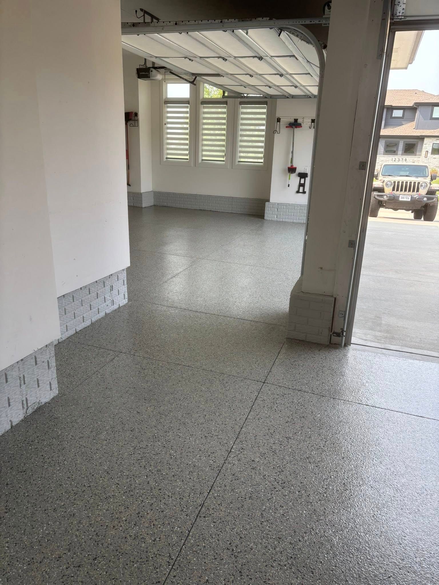 Garage interior with grey speckled flooring and matching baseboards, open door leading to a driveway and a parked vehicle.