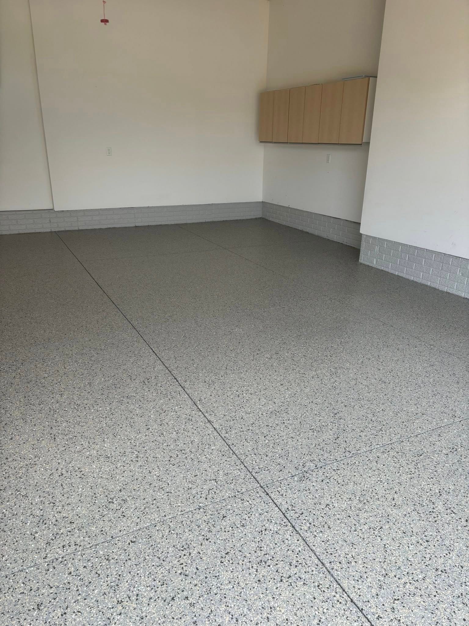 Empty garage interior with light grey, speckled epoxy flooring and matching baseboards against plain white walls.