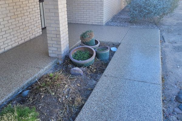A tan brick building entrance with a concrete walkway, a small dirt landscaping bed, and several empty green planters.