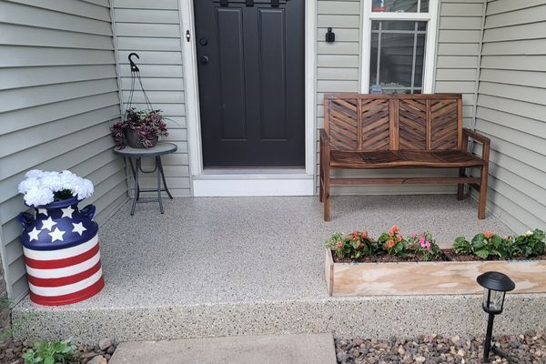 A front porch with a wooden bench, a flag-painted milk can with flowers, and a wooden planter box on a speckled floor.