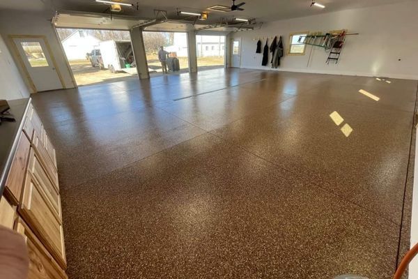 A bright, clean garage with a glossy, brown-speckled epoxy floor, open bay doors, and tools hanging on the white wall.
