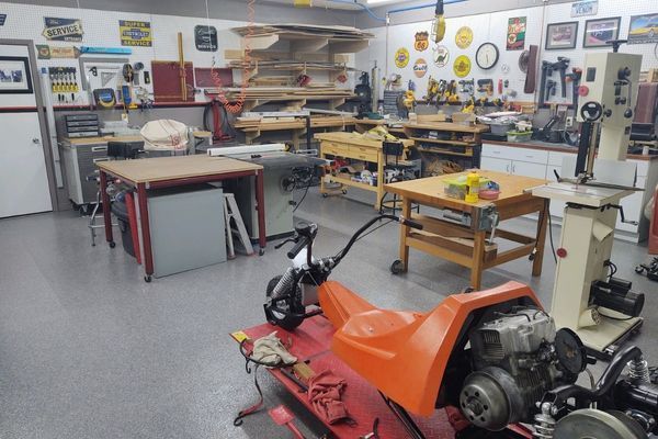 An orange motorized scooter sits on a red stand in a well-equipped workshop filled with tools and workbenches.