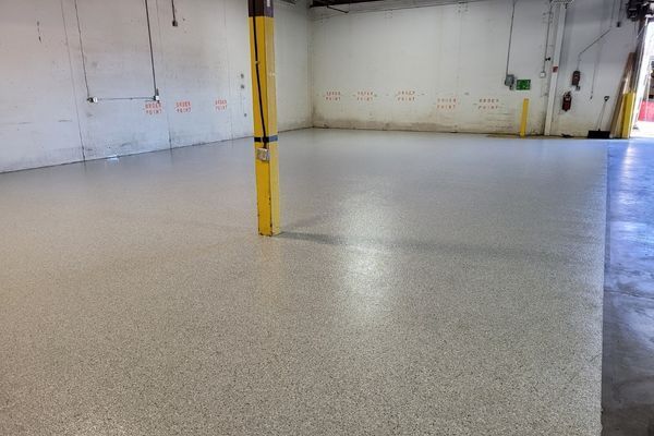 An empty industrial space featuring a light grey, speckled epoxy floor and a single yellow vertical support pole.