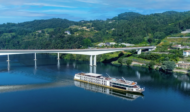 Navio de cruzeiro em um rio sob uma ponte, com colinas verdes ao fundo.