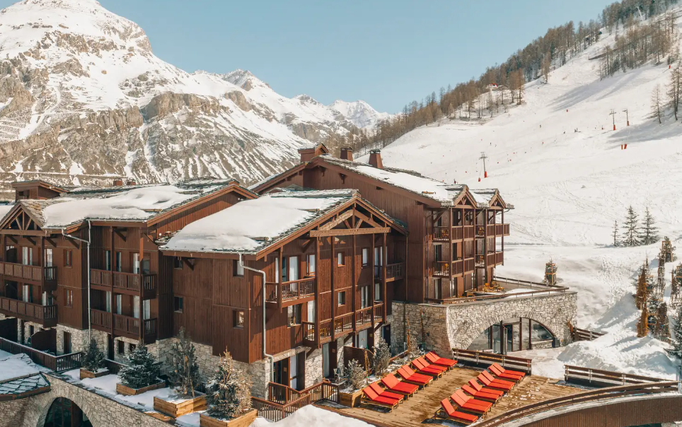 Chalé nevado na montanha com exterior de madeira, cadeiras de descanso vermelhas e montanhas ao fundo.