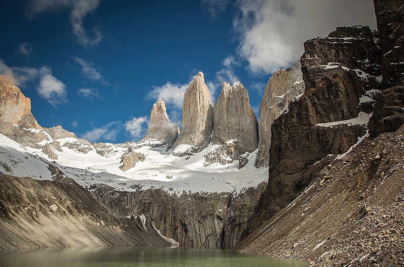 Picos nevados elevam-se sobre um lago glacial, emoldurados por encostas rochosas sob um céu azul.