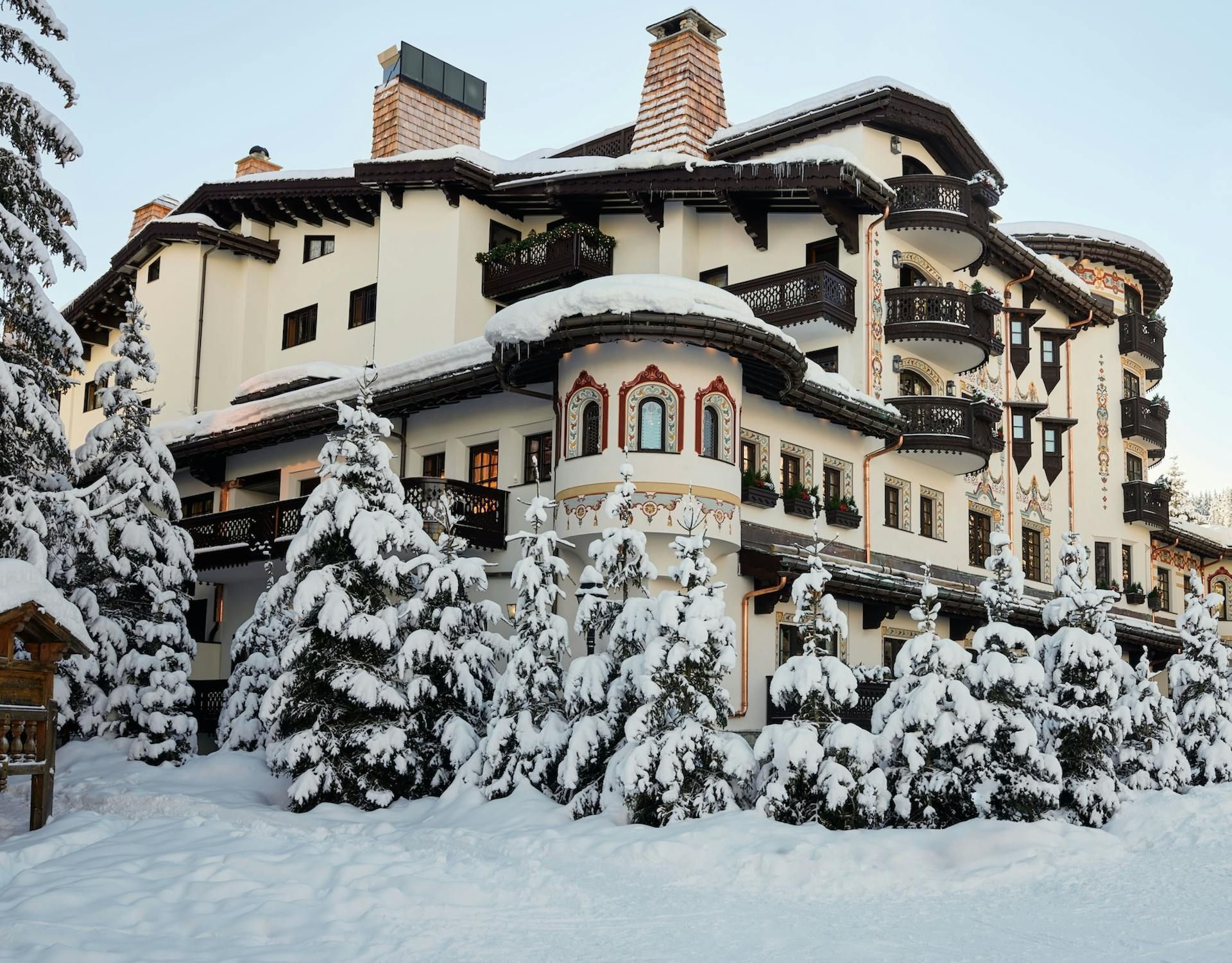 Edifício turístico coberto de neve com detalhes em arco, varandas e chaminés, rodeado por árvores perenes cobertas de neve.