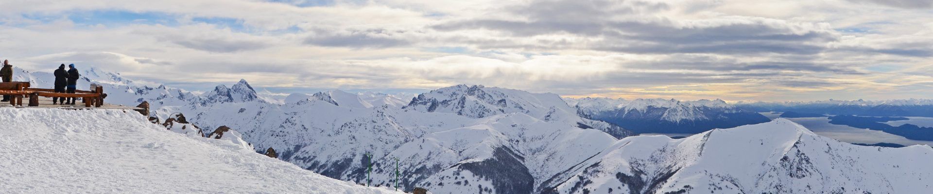Cordilheira coberta de neve sob céu nublado. Pessoas em uma plataforma de madeira com vista para a paisagem.