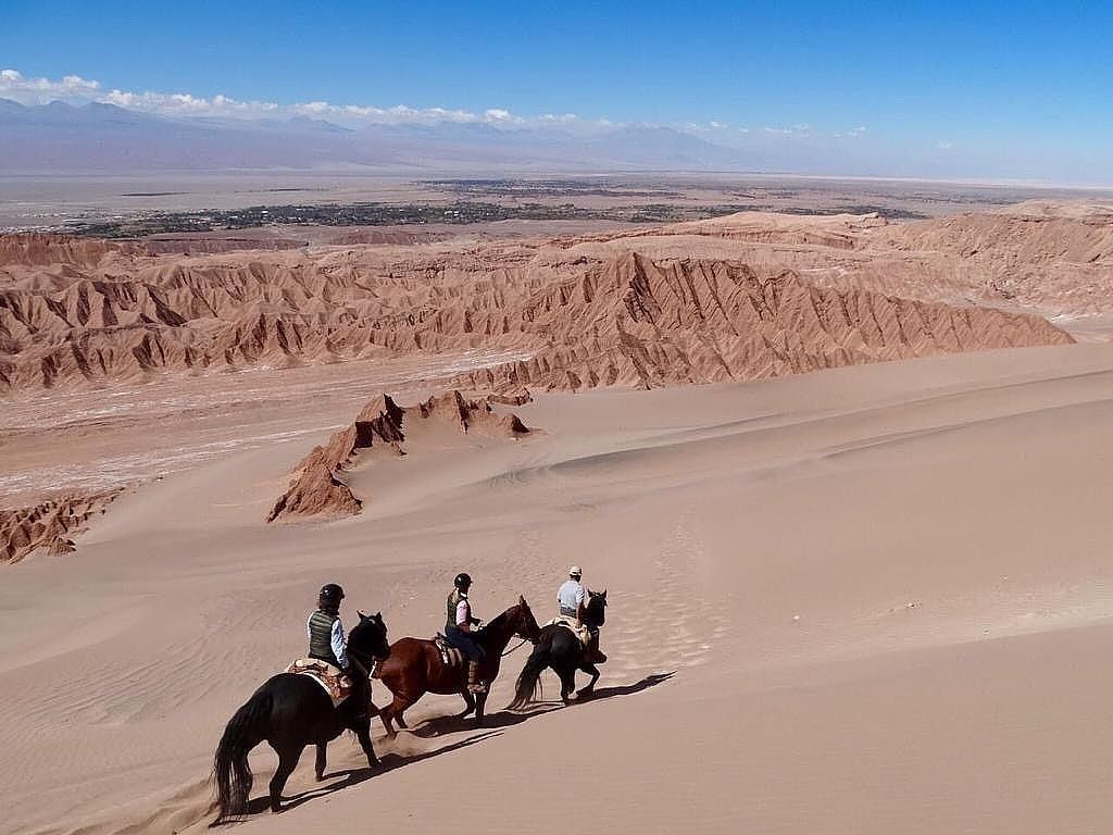 Pessoas cavalgam por uma paisagem desértica sob um céu azul.