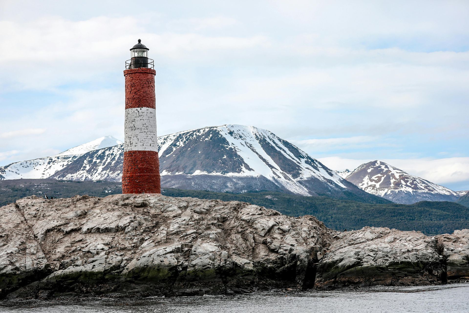 Farol listrado em vermelho e branco em uma ilha rochosa, com montanhas nevadas ao fundo.