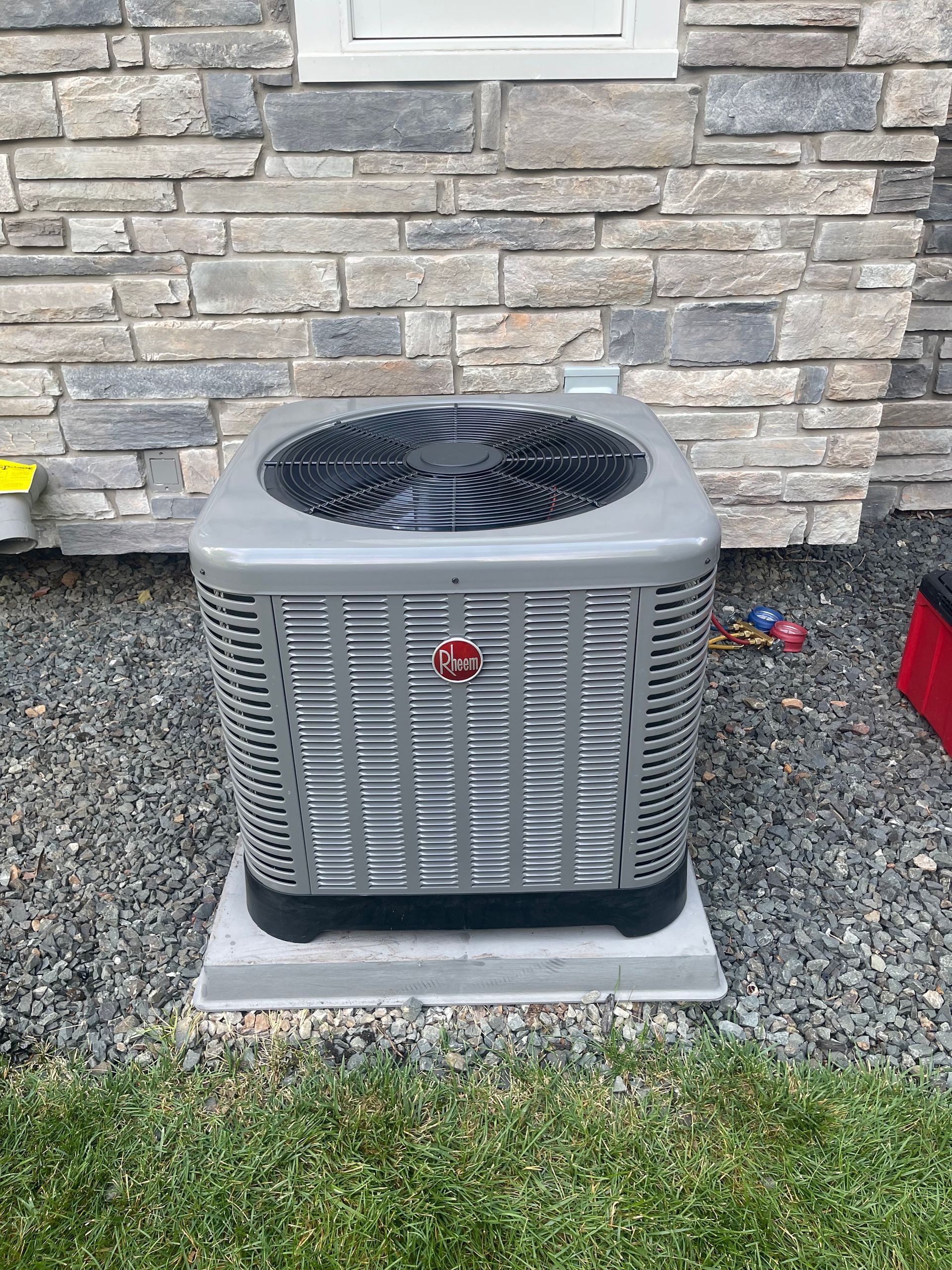 A gray air conditioner is sitting on a concrete base in front of a brick wall.