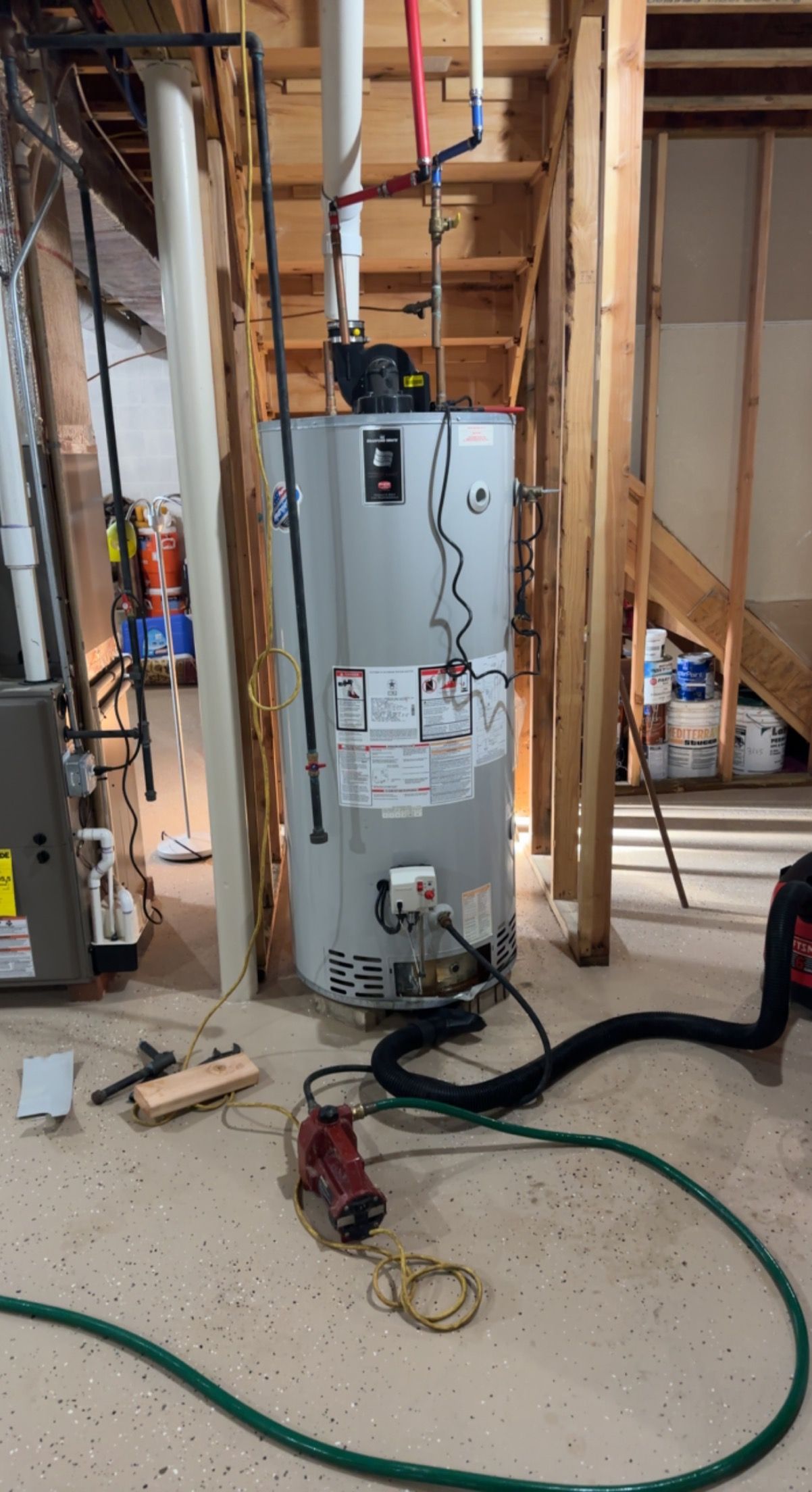A water heater is sitting in a basement next to a vacuum cleaner.