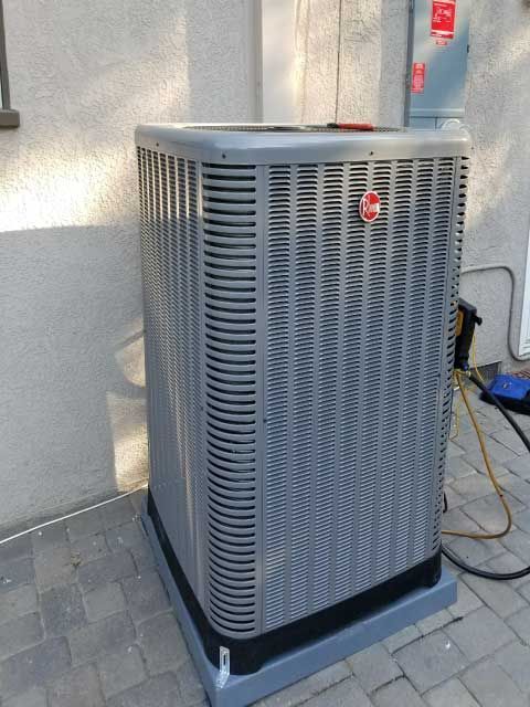 A gray air conditioner is sitting on a brick sidewalk in front of a building.