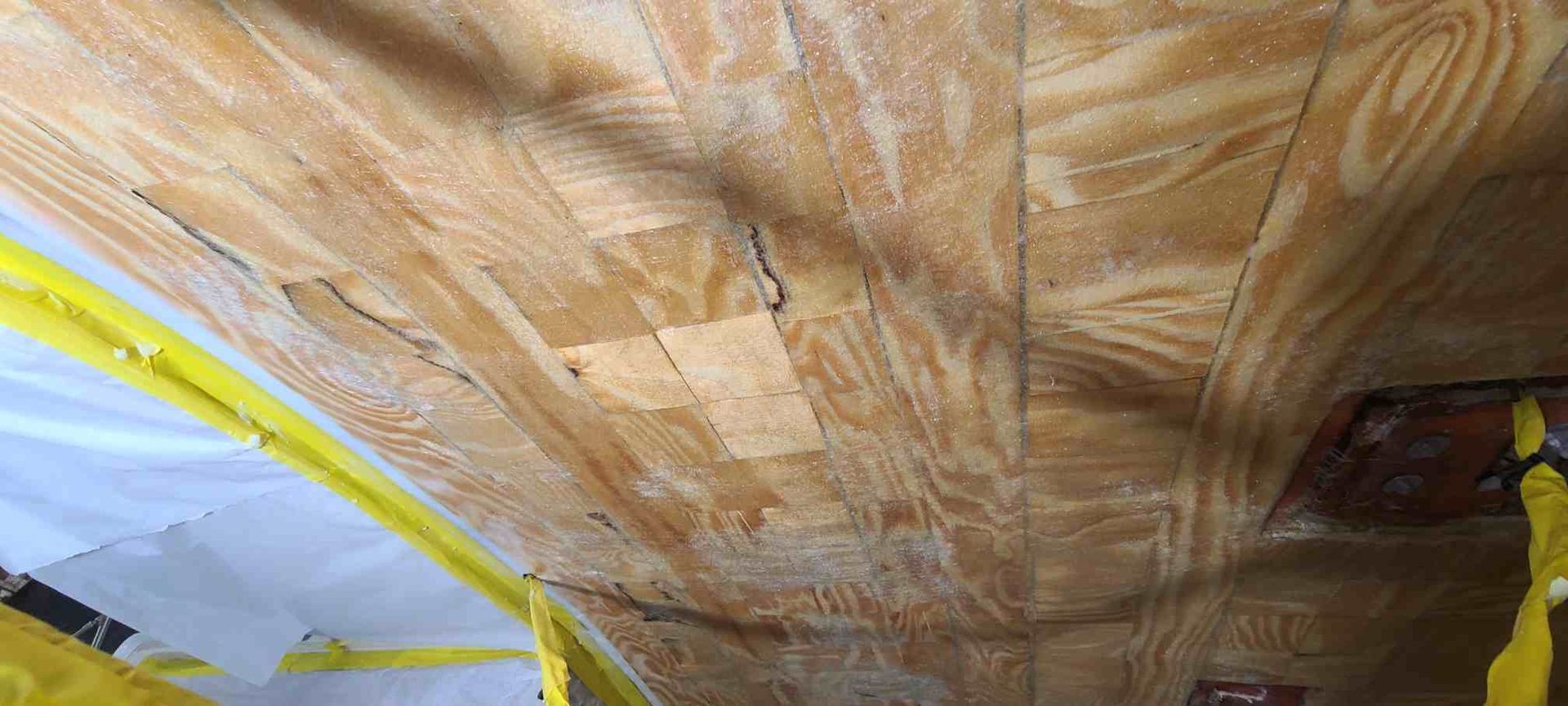 Wooden ceiling with cracks and some visible water damage. Yellow tape and plastic sheeting are visible.