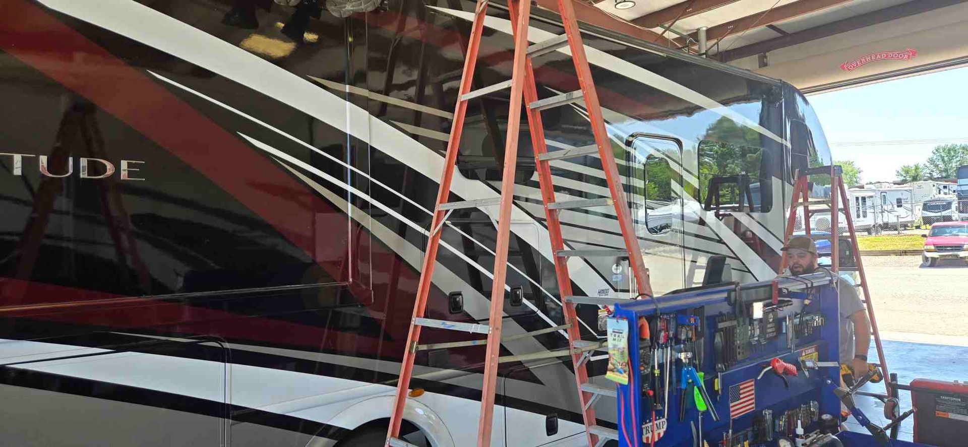 An RV parked inside a repair shop, with an orange ladder.
