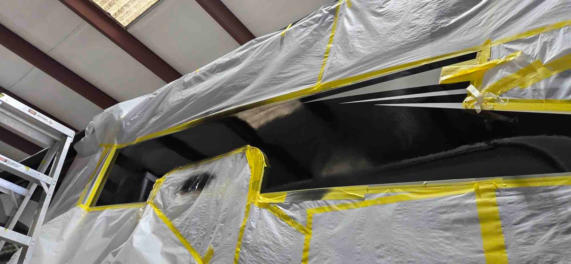 A vehicle window masked with plastic sheeting and yellow tape in a workshop, ready for painting.