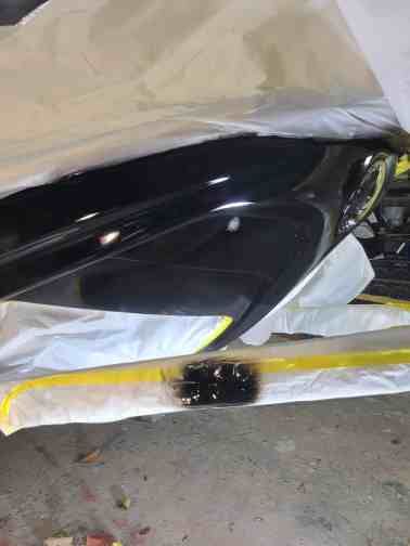 Black car bumper being painted, covered in white and yellow masking.