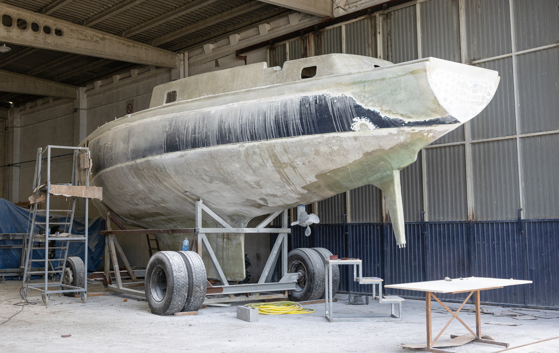 Sailboat hull on a wheeled cradle inside a workshop. Gray, with a black section on the bow.